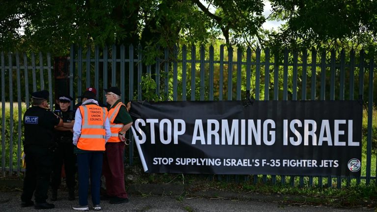 Protesters holding a large black banner that reads "STOP ARMING ISRAEL" near a metal fence with police officers nearby.