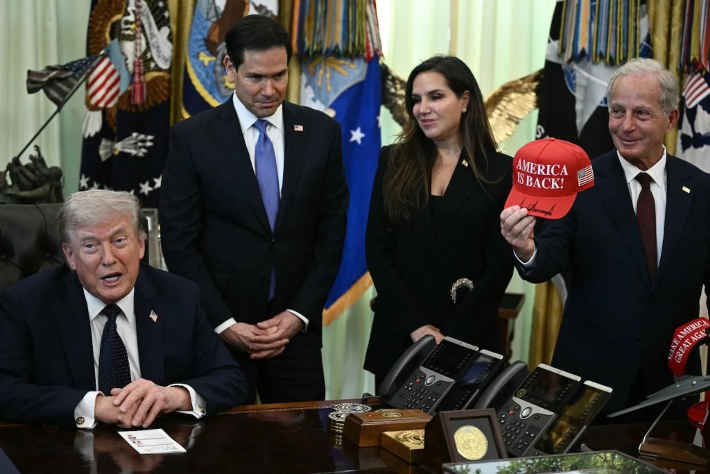 Donald Trump seated at a desk with advisors, including Marco Rubio, while an official holds up a red "America is Back!" hat.