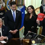 Donald Trump seated at a desk with advisors, including Marco Rubio, while an official holds up a red "America is Back!" hat.