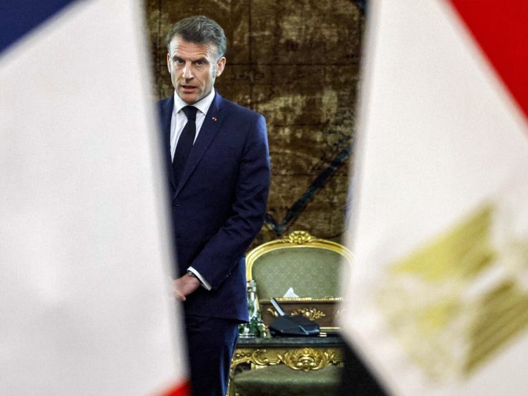 French President Emmanuel Macron standing in a formal room, flanked by partially visible flags, during a diplomatic engagement.