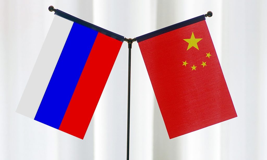 Small desktop flags of Russia and China standing crossed against a soft, light-colored background.