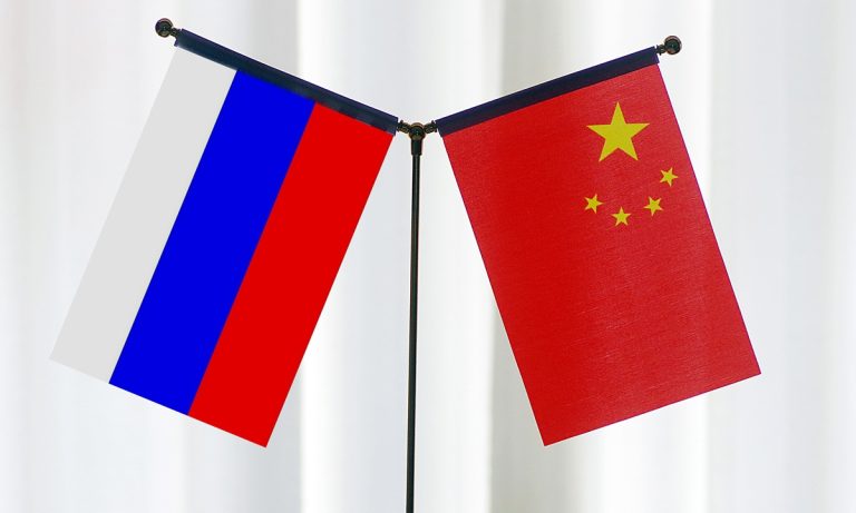 Small desktop flags of Russia and China standing crossed against a soft, light-colored background.