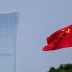 The Chinese national flag waving in the wind with a modern skyscraper in the background.
