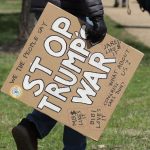 Person carrying a cardboard sign that reads "Stop Trump's War" at a protest.