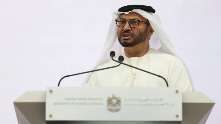 A man in traditional Emirati attire and glasses speaking behind a white podium featuring the United Arab Emirates Ministry of Foreign Affairs emblem.