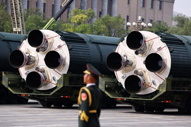 Large Chinese ballistic missile canisters on transport vehicles during a military parade with a soldier in the foreground.