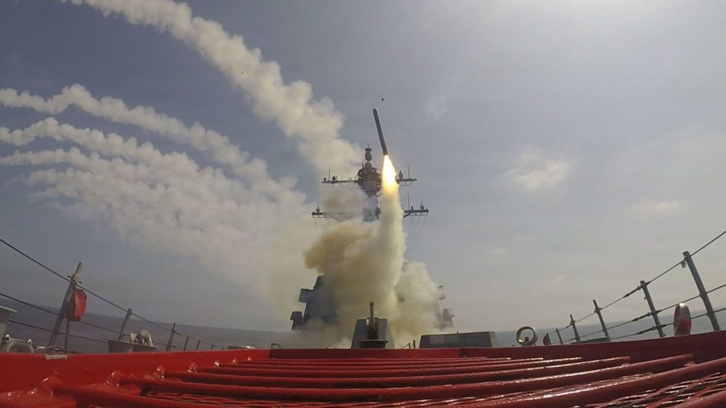 A missile being launched from the deck of a US Navy destroyer at sea, leaving a trail of white smoke.