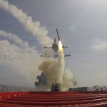 A missile being launched from the deck of a US Navy destroyer at sea, leaving a trail of white smoke.
