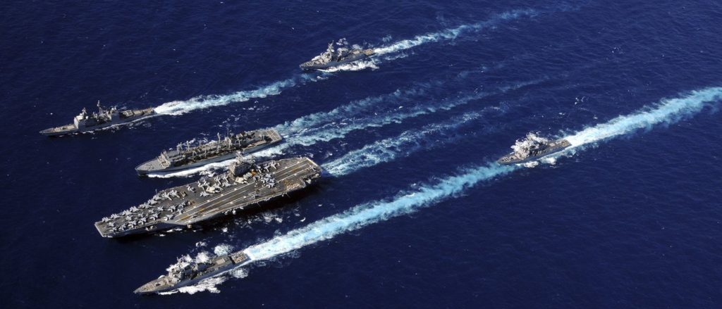 Aerial view of a US Navy carrier strike group moving across the blue ocean.