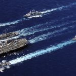 Aerial view of a US Navy carrier strike group moving across the blue ocean.