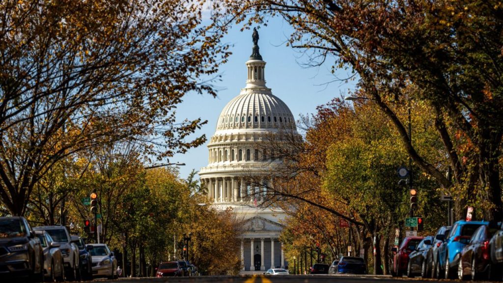 The United States Capitol building dome viewed from a tree-lined street with colorful autumn foliage and parked cars.