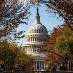 The United States Capitol building dome viewed from a tree-lined street with colorful autumn foliage and parked cars.