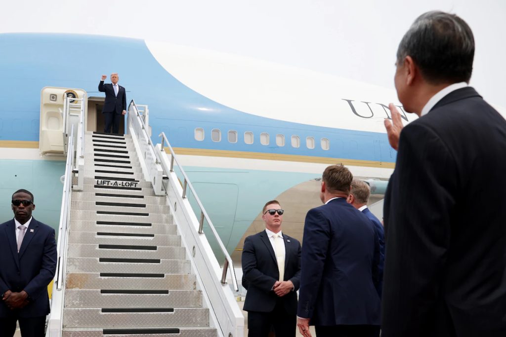 US President Donald Trump waving from the doorway of Air Force One while officials on the tarmac watch.