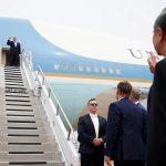 US President Donald Trump waving from the doorway of Air Force One while officials on the tarmac watch.