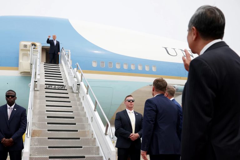 US President Donald Trump waving from the doorway of Air Force One while officials on the tarmac watch.