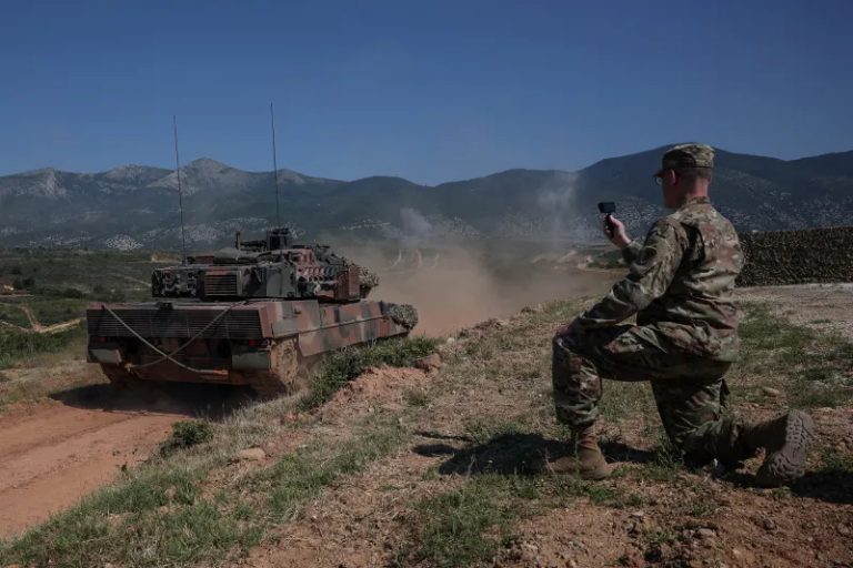 A soldier in camouflage kneeling on a dirt path while operating a drone controller, with a military tank nearby.