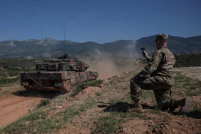 A soldier in camouflage kneeling on a dirt path while operating a drone controller, with a military tank nearby.