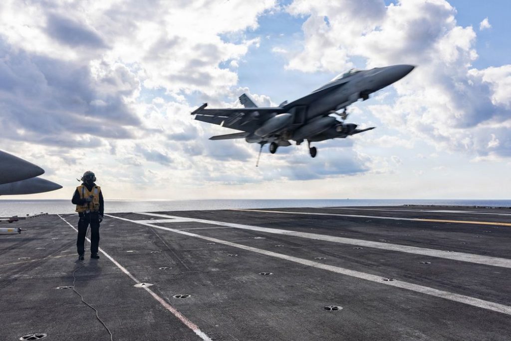 A US Navy F/A-18 Super Hornet fighter jet landing on the flight deck of an aircraft carrier.