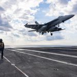 A US Navy F/A-18 Super Hornet fighter jet landing on the flight deck of an aircraft carrier.