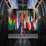 A person walking in front of a row of international flags set against a backdrop of server racks.
