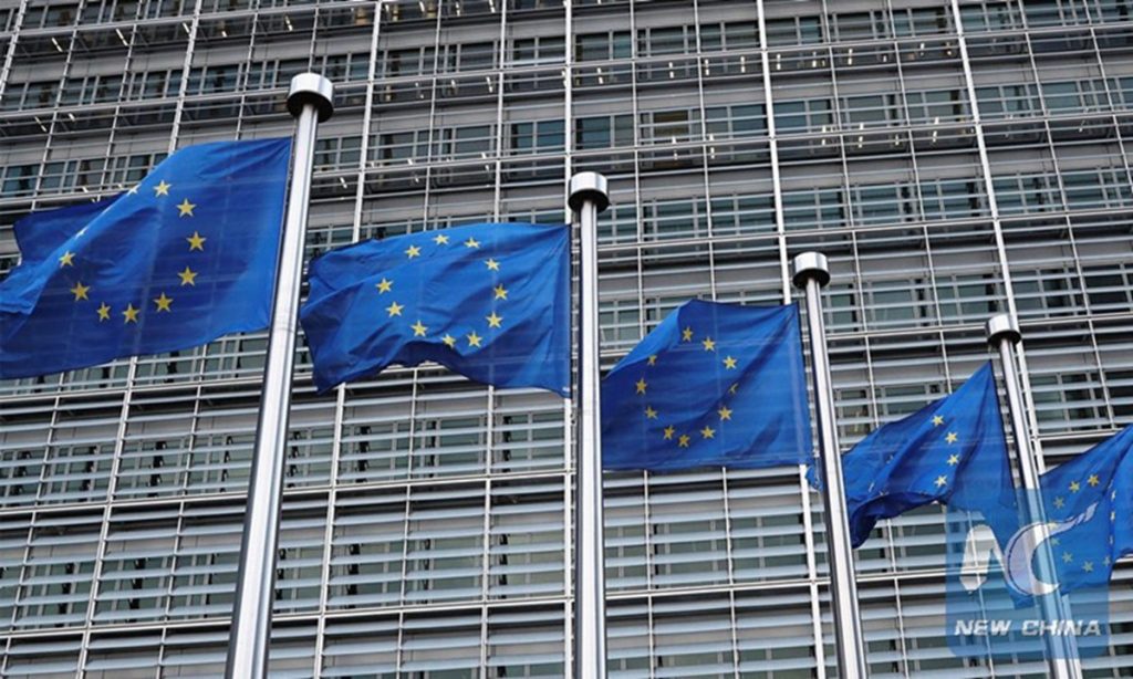 Row of European Union flags flying in front of the modern European Commission headquarters building.