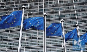 Row of European Union flags flying in front of the modern European Commission headquarters building.