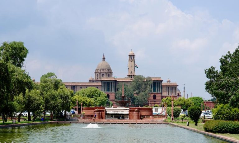 Exterior view of government buildings in New Delhi with greenery and a water fountain in the foreground.