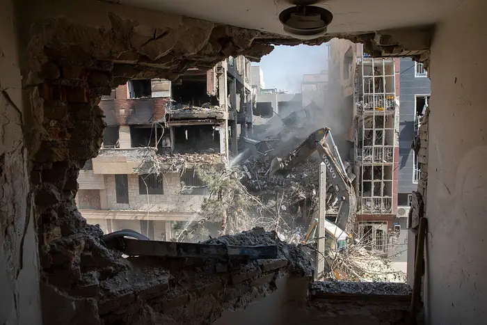 A construction vehicle clearing rubble from a damaged building as seen from inside a destroyed room.