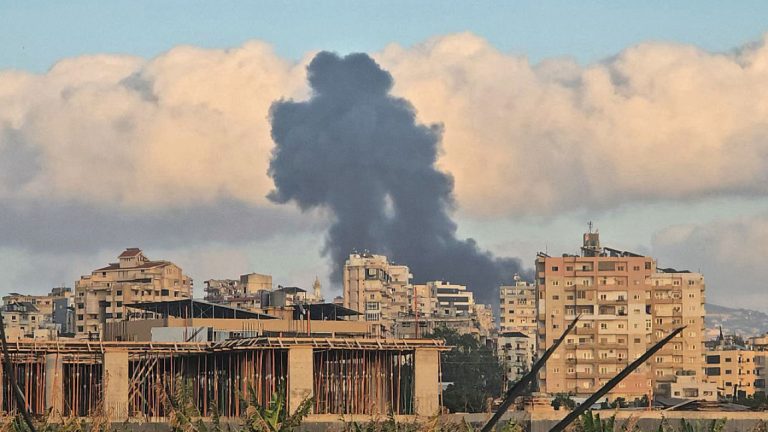 Thick black smoke rising into the sky over a Lebanese city with residential buildings in the foreground.