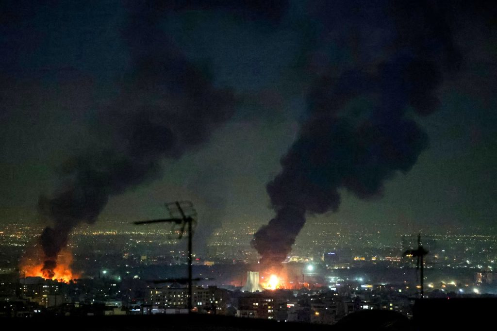 Night view of a city skyline with plumes of black smoke and fire caused by military conflict.