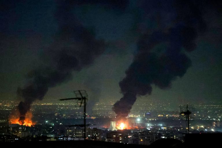 Night view of a city skyline with plumes of black smoke and fire caused by military conflict.