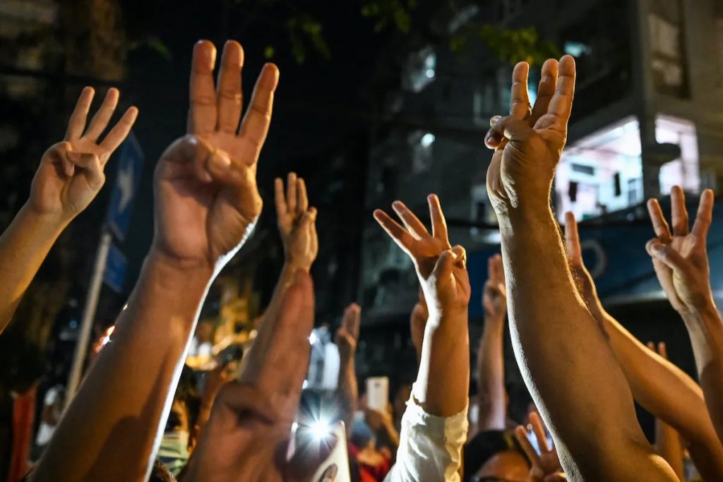 A crowd of protesters in Myanmar raising their hands in a three-finger salute at night.