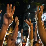 A crowd of protesters in Myanmar raising their hands in a three-finger salute at night.