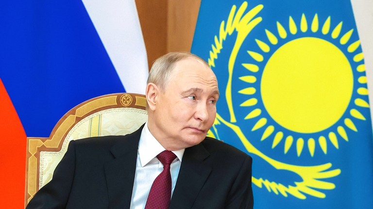 Vladimir Putin sitting in a chair in front of the Russian and Kazakhstani flags.