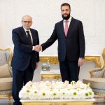 Two men in formal suits shaking hands in front of the flags of Lebanon and Syria.