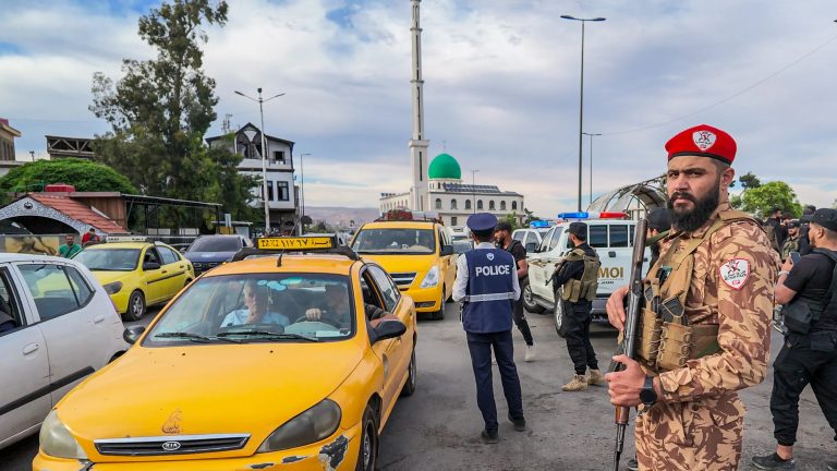 A soldier in military uniform standing guard near a mosque and city traffic.