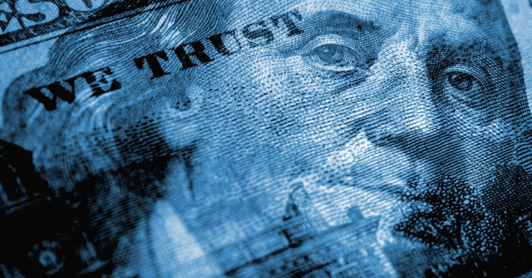 A close-up, blue-tinted macro shot of a United States one-hundred-dollar bill featuring the words "In God We Trust".