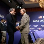 Two diplomats shaking hands at the Lake Lucerne Summit with flags of different nations displayed in the background.