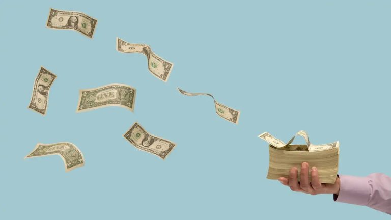 A hand holds a thick stack of U.S. dollar bills while other individual bills fly through the air against a light blue background.