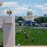 An aerial view of a green lawn in Washington, D.C., featuring a decorative archway and the U.S. Capitol building in the background.
