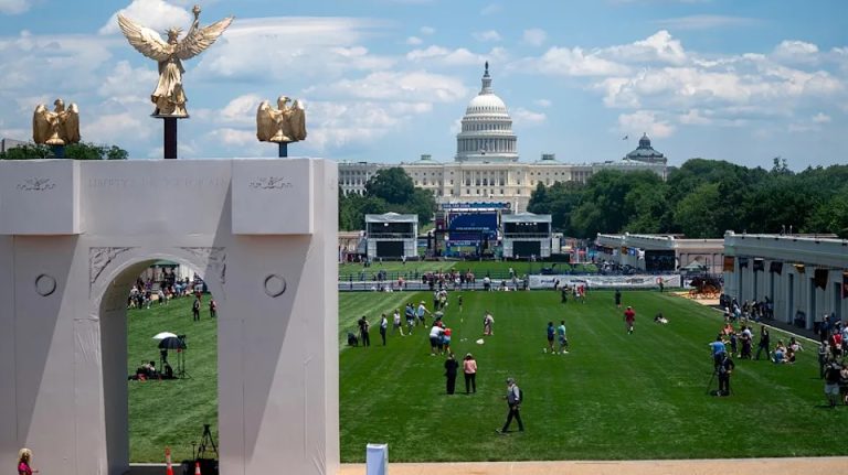 An aerial view of a green lawn in Washington, D.C., featuring a decorative archway and the U.S. Capitol building in the background.