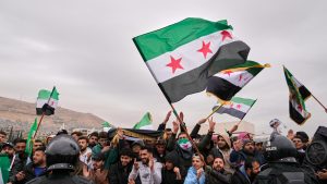 A crowd of protesters in Syria waving opposition flags in front of police officers.