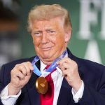 Donald Trump smiling while holding a gold medal around his neck.