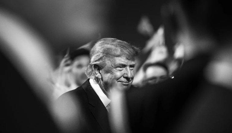 A black and white portrait of Donald Trump smiling among a crowd.