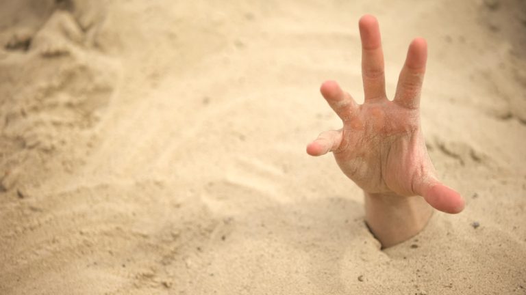 A human hand emerging from deep sand, reaching upward.