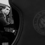 A man in a suit sitting at a table, viewed from behind a U.S. House of Representatives seal.