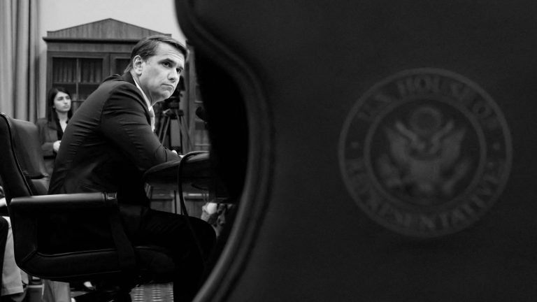 A man in a suit sitting at a table, viewed from behind a U.S. House of Representatives seal.