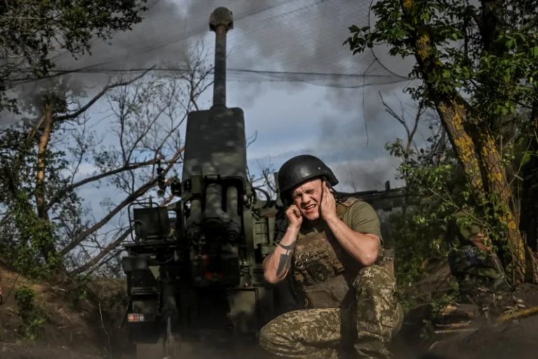 A Ukrainian soldier covering his ears near a heavy artillery piece during combat.