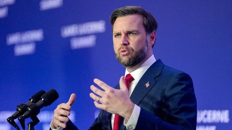J.D. Vance speaking at a podium in front of a blue background during a professional summit event.
