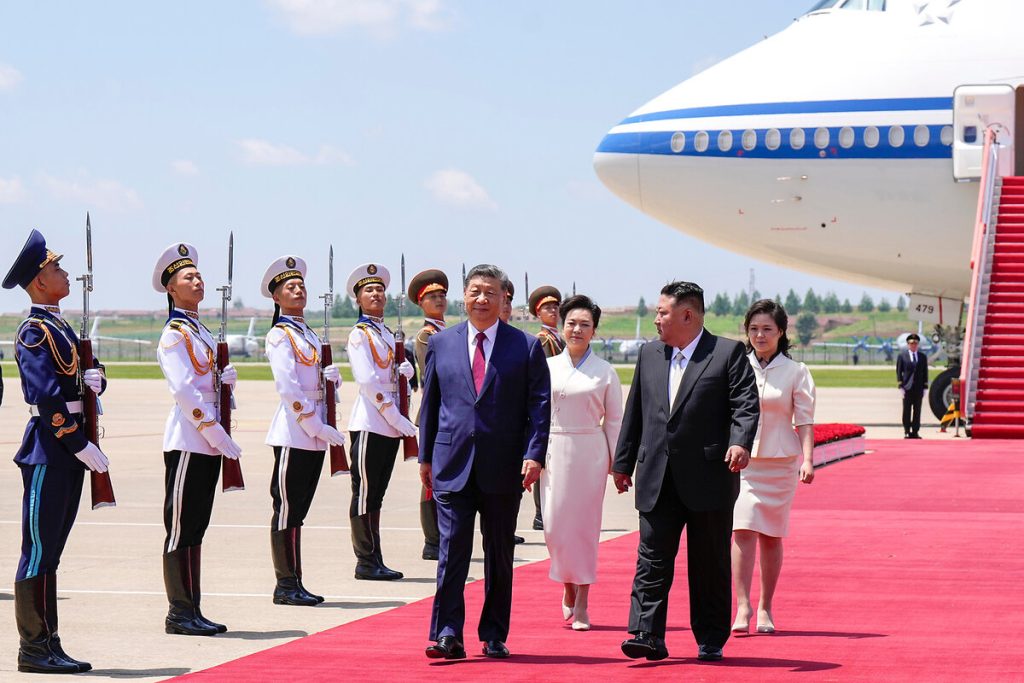 Xi Jinping and Kim Jong Un walking on a red carpet at an airfield with an honor guard and a large airplane in the background.